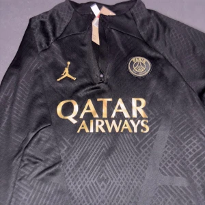 PSG x Jordan svart fotbollströja - Svart PSG fotbollströja från Jordan med guldfärgade detaljer och logotyper. Tröjan har halv dragkedja, diskret mönster i tyget och är tillverkad i ett lätt, sportigt material. Perfekt för dig som vill sticka ut på planen med stilren design och exklusiv känsla. Passar bra för M pris går alltid att diskutera