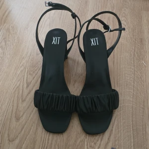 Svarta sandaler med klack från XIT strl 40 - Stilrena svarta sandaler från XIT med fyrkantig tå, bred rynkad rem över foten och smala ankelband med spänne. Skorna har en stabil blockklack och är tillverkade i syntetmaterial. Endast använda en kväll då I stort sett nya