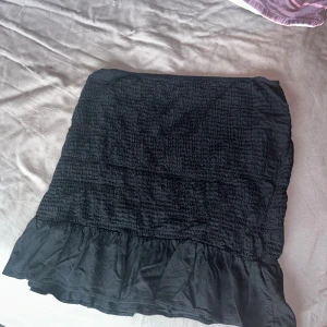 Svart volangkjol från BikBok - Supersöt svart kortkjol från BikBok med smockad struktur och volangkant nertill. Kjolen är elastisk i midjan och har en lätt och luftig känsla, perfekt för att styla med en crop top eller oversized hoodie.