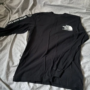 Svart långärmad tröja The North Face - Svart långärmad tröja från The North Face med vit logga och koordinater tryckta på bröstet. Texten 'Never Stop Exploring' och 'Since 1966' finns på ärmarna. Tröjan är i mjuk bomull och har normal passform med ribbade muddar vid ärmslut.