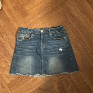 Blå jeanskjol med rå kant - Snygg blå jeanskjol med slitna detaljer och rå, fransig nederkant. Klassisk femficksmodell med knapp och dragkedja framtill. Perfekt för dig som gillar en avslappnad och trendig stil.