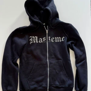 Svart hoodie från Masteme med dragkedja - Svart hoodie från Masteme med stor huva och dragkedja framtill. Tröjan har vita bokstäver i gotisk stil över bröstet och två stora fickor framtill. Perfekt för en avslappnad streetwear-look.
