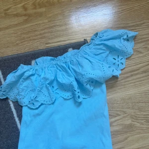 Ljusblå offshoulder topp med volang - Superfin ljusblå offshoulder topp med bred volangkant och broderade blommor. Toppen är ribbad i tyget och har en luftig känsla, perfekt för varma dagar. Volangen har fina håldetaljer som ger en söt och somrig look.