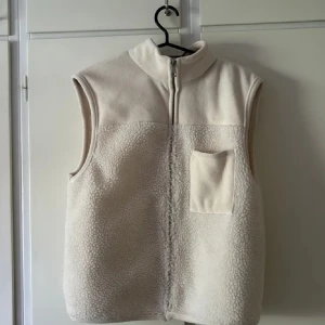 Beige fleeceväst med dragkedja - Mysig beige fleeceväst med hög krage och hel dragkedja framtill. Västen har en enkel design med en bröstficka på vänster sida och mjukt, fluffigt material som ger en chill vibe. Perfekt att slänga på över en hoodie när det är lite kyligt ute.