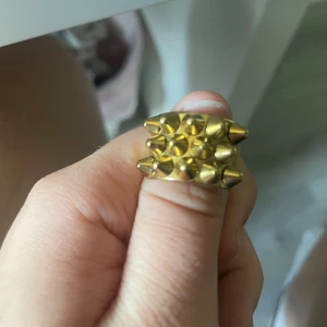 Guldig ring med nitar - Cool och chunky ring i guldtonad metall med flera spetsiga nitar på ovansidan. Ringen har en bred och fyrkantig form som verkligen sticker ut och ger en edgy vibe. Perfekt accessoar för dig som vill ha något unikt och vågat i din stil. storlek L men passar även M/S