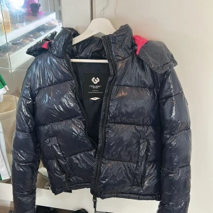 Mörkblå pufferjacka - Säljer en glansig mörkblå pufferjacka från Ragwear i storlek XS. Jackan har huva, dragkedja och en cool detalj med rosa inslag på axeln. Perfekt för kalla dagar och riktigt streetig look.