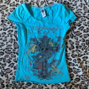 Ed Hardy t-shirt  - Blå t-shirt från Ed Hardy med stort färgglatt tryck och rhinestones framtill. Klassisk rund halsringning och korta ärmar. Materialet är mjuk bomull som känns skönt mot huden. Tight men stretchigt tyg.
