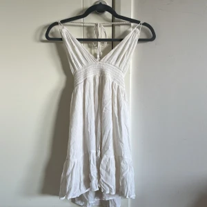 Vit somrig kortklänning med volang - Superfin vit kortklänning i luftig bomull med volangkant och smock upptill. Klänningen har djup v-ringning, knyt i nacken och öppen rygg. Perfekt för varma dagar och ger en avslappnad, bohemisk vibe.