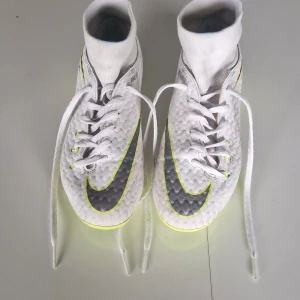 Nike Hypervenom vita fotbollsskor 33.5 - Nike Hypervenom fotbollsskor i vitt med neongula detaljer och svart swoosh. Skorna har snörning, hög strumpkrage och plös, samt platt sula med dobbar för konstgräs. Ovandelen är i syntetmaterial med textur för bättre bollkontroll. Perfekta för dig som vill sticka ut på planen. Skorna går över 1000 nya.