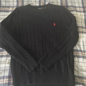 Svart kabelstickad tröja Polo Ralph Lauren - Svart kabelstickad tröja från Polo Ralph Lauren med klassisk röd broderad logga på bröstet. Tröjan har rund halsringning, långa ärmar och ribbade muddar. Perfekt för dig som gillar en stilren och tidlös look. Den är helt ny passa tyvärr inte mig 