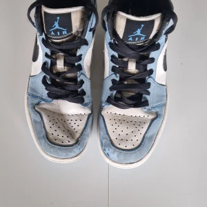 Nike Air Jordan 1 Mid UNC blå/vit storlek 38 - Nike Air Jordan 1 Mid i UNC-färg med ljusblå och vita paneler, svarta detaljer och snörning. Klassisk Jordan-logga på sidan och tungans etikett. Ovandel i skinn och mocka, perforerad tåbox och svart swoosh. Perfekta för dig som gillar streetstyle och sneakers med ikonisk design.