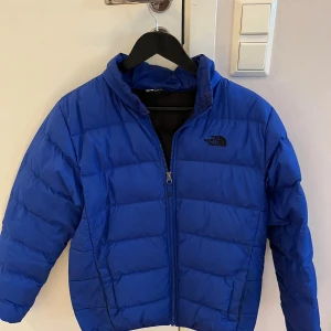 Blå pufferjacka från The North Face - Blå pufferjacka från The North Face med svart logga på bröstet och ryggen. Jackan har dragkedja framtill, hög krage och är quiltad för extra värme. Perfekt för kalla dagar och har en clean, sportig look. Jackan sitter stort på mig som är 175 cm.