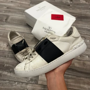 Valentino Garavani Open Sneakers vit/svart - Snygga Valentino Garavani Open sneakers i vitt kalvskinn med en bred svart läderdetalj över mitten. Klassisk rund tå, platt vit sula och diskreta nitar baktill. Perfekta för dig som gillar lyxiga och stilrena sneakers med en unik touch.