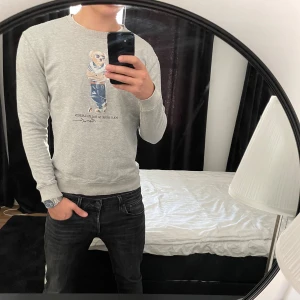 Grå sweatshirt Polo Bear Ralph Lauren - Säljer en grå sweatshirt från Polo Ralph Lauren med ikoniskt Polo Bear-tryck på bröstet. Tröjan har rund hals, långa ärmar och är tillverkad i mjuk bomullsmix. Perfekt för dig som gillar klassisk och avslappnad stil.