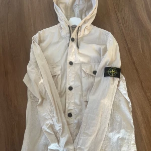 Beige vindjacka från Stone Island - Snygg beige vindjacka från Stone Island med huva och svarta knappar. Jackan har två stora bröstfickor med lock och den klassiska loggan på vänster ärm. Lätt material och dragsko i huvan gör den perfekt för vår och höst. 