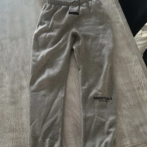 Grå mjukisbyxor Essentials Fear of God - Grå mjukisbyxor från Essentials Fear of God med svart logga på benet. Byxorna har elastisk midja med snörning och muddar vid bensluten. Tillverkade i mjukt bomullsmaterial för en chill och avslappnad look. Använt ett fåtal gånger 
