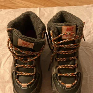 Nike City Classic Premium boots - Nike City Classic Premium boots i mörkgrönt skinn med grov beige sula och orange-mönstrade snören. Insidan är fodrad med mjukt grått fleecefoder för extra värme. Perfekta för kalla dagar med robust design och snygga detaljer.