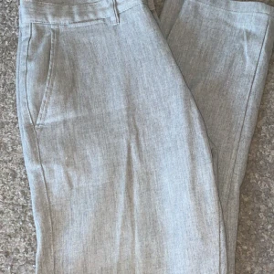 Ljusgrå kostymbyxor med knapp - Snygga ljusgrå kostymbyxor med klassisk passform och diskret melerad yta. Byxorna har hällor för bälte, sidfickor och stängs med knapp och dragkedja. Perfekta för en clean och stilren look.