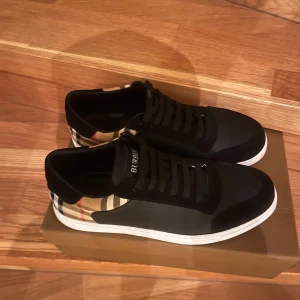 Burberry skor - Snygga sneakers från Burberry med klassiskt rutigt mönster i beige och svart på hälen. Ovandelen är svart med vita sulor och svarta snören. Perfekta för dig som vill ha en stilren men ändå unik look.