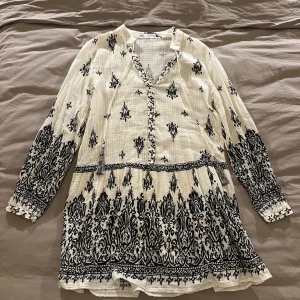 Vit och svart mönstrad klänning från Zara - Supersöt kortklänning från Zara i vitt med svart broderat mönster. Klänningen har långa ärmar, v-ringning med knyt och volang nedtill. Perfekt för sommarens alla tillfällen och ger en bohemisk vibe.