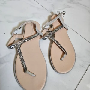 Silvriga sandaler med strassdetaljer - Snygga sandaler i ljusbeige med tunna remmar och glittrande strassdetaljer längs remmarna. Perfekta för sommardagar och har en elegant design med öppen tå och spänne vid ankeln.