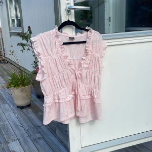 Rosa volangblus från Gina Tricot - Supersöt ljusrosa blus från Gina Tricot med volanger och knytning framtill. Blusen är ärmlös och har ett lätt strukturerat tyg med diskret mönster. Perfekt för en somrig look och passar till både jeans och kjol.