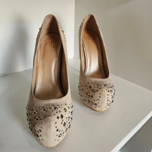 Beige pumps med strass från NLY - Snygga beige pumps från NLY med höga klackar och spetsig tå. Skorna är täckta med glittriga strass-stenar som ger en riktigt festlig vibe. Tillverkade i mockaimitation och kommer med extra klackpluppar. Perfekta för dig som vill sticka ut.