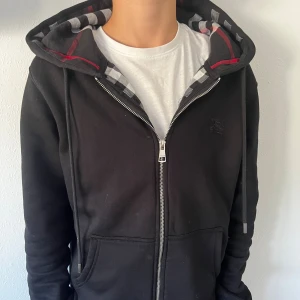 Svart Burberry hoodie med rutig luva - Svart hoodie från Burberry i storlek M med dragkedja framtill och klassiskt rutig foder i huvan i svart, vitt och rött. Tröjan har huva med snörning, två fickor och diskret Burberry-logga på bröstet. Perfekt för dig som gillar stilrena och exklusiva plagg.
