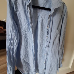 Ljusblå linneskjorta från H&M - Säljer en ljusblå skjorta från H&M i linnemix. Skjortan har klassisk krage, knäppning framtill och en bröstficka. Långärmad och med avslappnad passform, perfekt för en chill och stilren look.