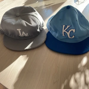 Två New Era kepsar LA & KC MLB - Säljer två klassiska New Era kepsar med broderade loggor: en grå LA Dodgers med vit logga och en ljusblå Kansas City Royals med blå skärm och vit KC-logga. Båda är i basebollstil och har MLB-märke bak. Perfekta för dig som gillar sportig streetstyle.