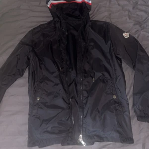 Svart vindjacka från Moncler - Svart vindjacka från Moncler med huva som har röd, vit och blå rand längs kanten. Jackan har dragkedja framtill, två fickor med tryckknappar och Moncler-logga på vänster ärm. Lätt och smidig modell i syntetmaterial, perfekt för blåsiga dagar.