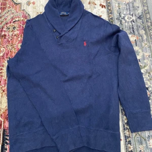 Tröja från Polo Ralph Lauren - Säljer en marinblå långärmad tröja från Polo Ralph Lauren med snygg sjalkrage och två knappar vid halsen. Tröjan har röd broderad logga på bröstet och är gjord i mjuk bomull.