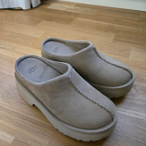 Beige platåtofflor från UGG, stl 36 - Säljer ett par UGG New Height Clogs i storlek 36.  Färg: klassisk beige/brun (UGG’s signaturlook).  Använda mycket sparsamt – i nästintill nyskick.  Bekväma med träklack och fårskinnsdetaljer som gör dem både trendiga och sköna.  Passar perfekt nu till höst/vinter för en stilren men avslappnad look.   💰 Pris: 1000kr 