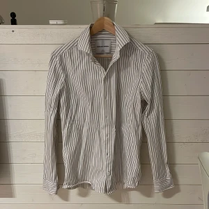 Vit randig skjorta Shaping New Tomorrow - Vit skjorta med svarta tunna ränder från Shaping New Tomorrow. Slim fit-modell med klassisk krage och knäppning framtill. Långärmad och riktigt clean look som passar till många olika outfits.