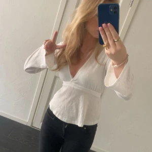 Vit omlottblus med v-ringning - Säljer en vit blus med omlottkänsla och djup v-ringning. Toppen har långa, vida ärmar och markerad midja som ger en snygg siluett. Perfekt att styla med jeans eller kjol för en fräsch look. Man knyter själv i ryggen. Org pris 300