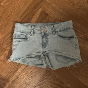 Ljusblå jeansshorts från Nelly - Säljer ett par ljusblå jeansshorts från Nelly med slitna detaljer och råa kanter. Shortsen har klassisk femficksdesign, bälteshällor och snygga röda sömmar vid fickorna samt broderade bakfickor. Perfekta för varma dagar och en avslappnad look.