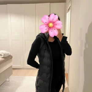 Svart lång dunväst med huva - Snygg svart lång dunväst med huva och dragsko. Västen har quiltad design, dragkedja framtill och två fickor på sidorna. Perfekt att slänga över en hoodie för en chill look köpt på zara helt ny.