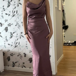 Plommonlila långklänning från Adoore  - Säljer en elegant plommonlila långklänning från Adoore i satinliknande material. Klänningen har draperad halsringning, figurnära passform och är ärmlös med snygg rynkdetalj i sidan. Perfekt för dig som vill sticka ut med en stilren och trendig look.