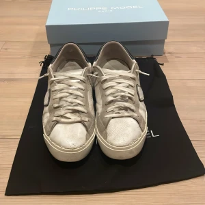 Philippe Model vita sneakers i skinn - Snygga vita sneakers från Philippe Model i skinn med klassisk låg profil och svarta detaljer vid hälen. Skorna har snörning, rund tå och en platt sula. Perfekta för dig som gillar stilrena skor.Skorna är i okej skick med slitningar under hälen 