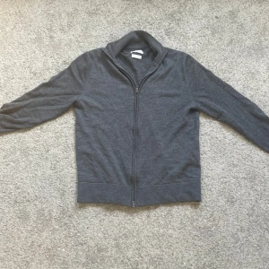 Grå merinoull kofta från These Glory Days - Mörkgrå kofta i 100% merinoull från These Glory Days. Klassisk modell med hel dragkedja framtill och ståkrage. Perfekt för lager-på-lager och riktigt skön mot huden. Enkel och stilren design som funkar till det mesta.