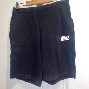 Svarta Nike shorts i bomull - Svarta shorts från Nike med vit logga på vänster ben. Mjukt bomullsmaterial och elastisk midja för skön passform. Klassisk design med sidofickor och en bakficka. Perfekta för chill dagar eller träning.