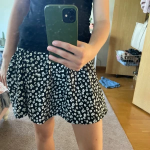 Svart blommig kortkjol - Supersöt svart kortkjol med vita blommor i ett tätt mönster. Kjolen har en lätt utställd form och är perfekt för dig som gillar en lekfull och trendig stil. Passar perfekt till en enkel topp eller hoodie.