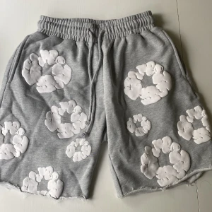 Denim Tears Grå färg  - Grå shorts från Denim Tears med vita, upphöjda blommönster över hela plagget. Modellen har elastisk midja med snörning, råa kanter och en bakficka. Mjukt bomullsmaterial som är skönt mot huden och ger en chill vibe.