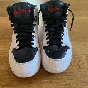 Nike Air Jordan 1 svartvita sneakers - Snygga Nike Air Jordan 1 sneakers i svart och vitt skinn med svarta snören och röd Jumpman-logga på plösen och sidan. Klassisk rund tå, perforerade detaljer och platt sula. Perfekta för dig som gillar streetstyle och vill sticka ut med ikoniska sneakers.