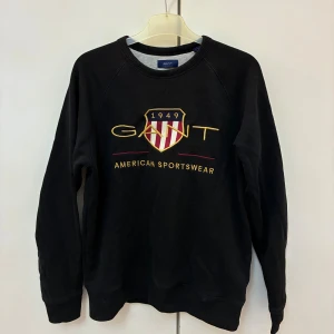 Svart sweatshirt från GANT med brodyr - Svart sweatshirt från GANT med broderad logga och sköld på bröstet i gult, rött och vitt. Klassisk rund halsringning och långa ärmar. Tillverkad i mjuk bomullsmix. Sitter som Storlek S/XS. Köpt för 800kr. 