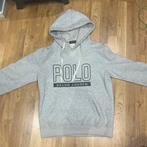 Grå hoodie från Polo Ralph Lauren - Säljer en grå hoodie från Polo Ralph Lauren med stor logga framtill. Tröjan har huva med snörning och en klassisk känguruficka. Perfekt för dig som gillar en enkel och stilren look. Materialet är mjukt och skönt, passar till chill dagar.