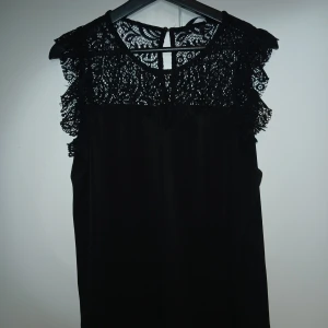 Svart blus med spetsdetaljer - Supersnygg svart blus med transparent spets upptill och volangdetaljer vid axlarna. Ärmlös modell som ger en elegant och trendig look. Perfekt för dig som gillar stilrena plagg med det lilla extra. Tillhörande shorts medföljer.