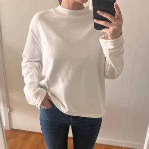 Vit loose sweatshirt med hög krage - Enkel och stilren vit sweatshirt med loose passform och hög ribbad krage. Tröjan har långa ärmar och är gjord i mjuk bomullsmix, perfekt för en chill och avslappnad look. Passar dig som gillar minimalistisk stil och vill ha något bekvämt till vardags.