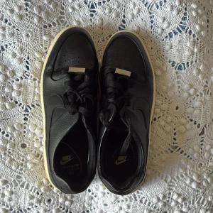 Nike Air Force 1 svarta sneakers från Nike - Svarta Nike Air Force 1 sneakers i skinn med vit, tjock platt sula och klassisk snörning. Swoosh-logga på sidan och AF-1 broderat bak. Stilrena och passar perfekt till streetwear. Perforerad tå för extra ventilation. Använd en gång, men inga slitage. Storlek 40.