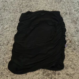 Svart kortkjol med rynk från SHEIN - Svart kortkjol från SHEIN i stretchigt syntetmaterial. Kjolen har snygga rynkade detaljer längs sidorna som ger en cool och trendig look. Perfekt för dig som gillar en enkel men ändå stilren vibe.